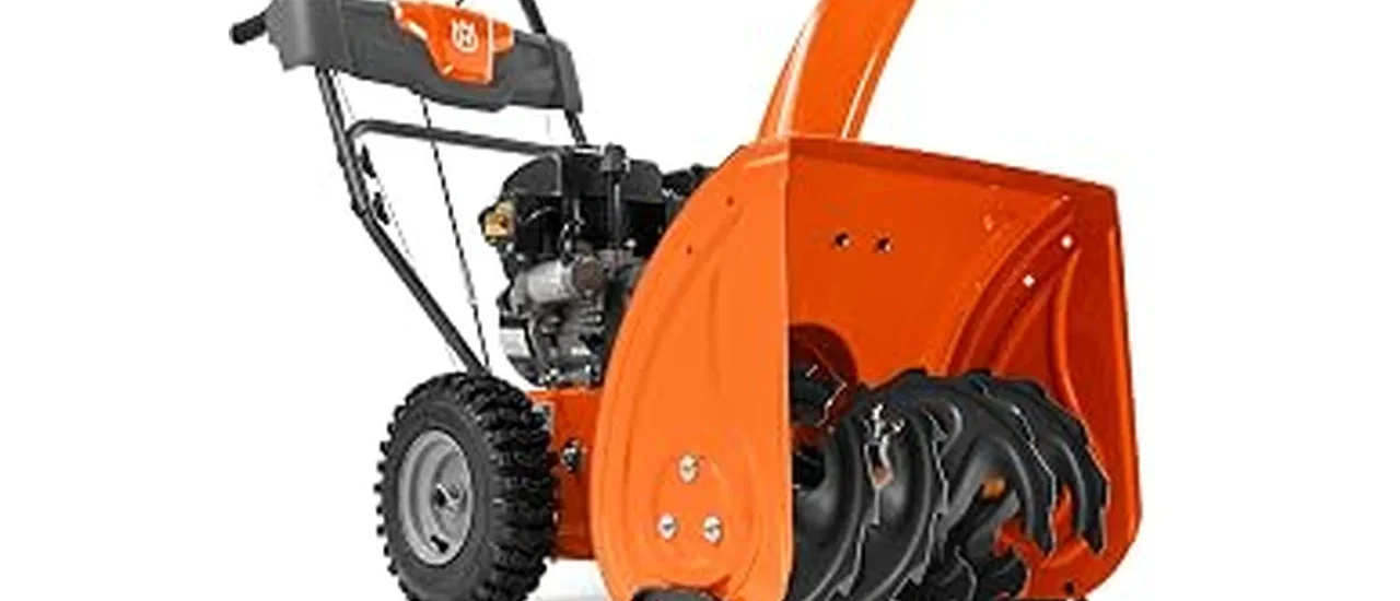 The image shows a powerful two-stage snow blower clearing a large, snow-covered driveway.