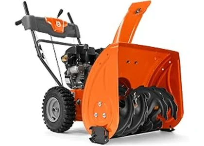 The image shows a powerful two-stage snow blower clearing a large, snow-covered driveway.